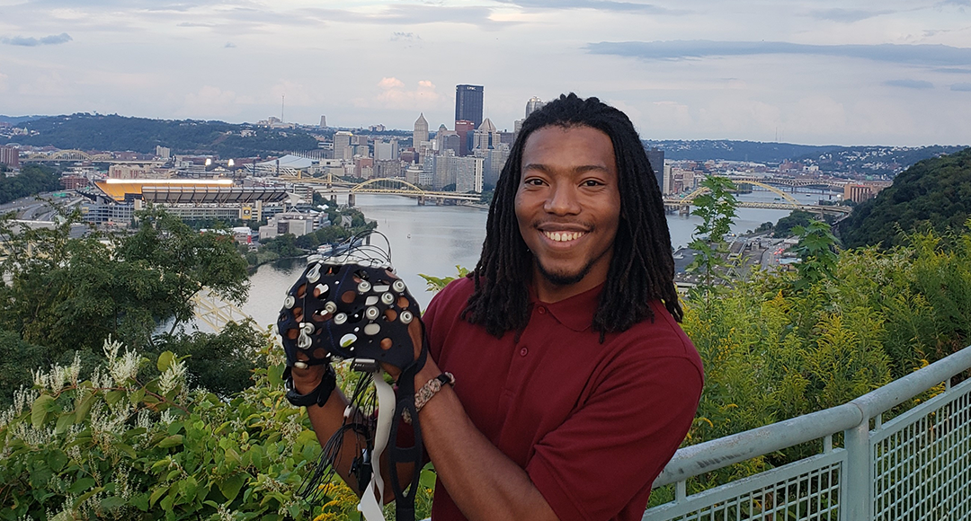 Student holding a black cap with multiple silver nodes on it at Mt. Washington, an overlook right outside of Pittsburgh.