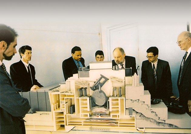 Seven men in a room with a model of a city on the table in front of them.