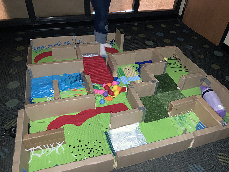 A floor maze with someone walking through with socks on. There are balloons, legos, and other items you wouldn't want to step on in addition to soft places through the maze where you can step.