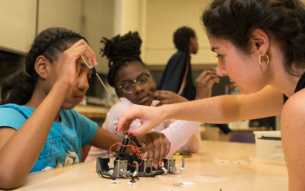 Female college student working on a tabletop robot with two young girls
