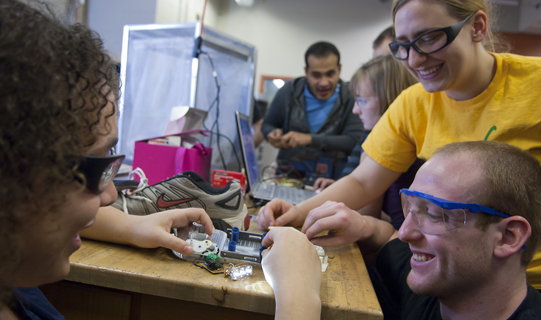 Three smiling students building an electronic circuit in a lab with other students in the background