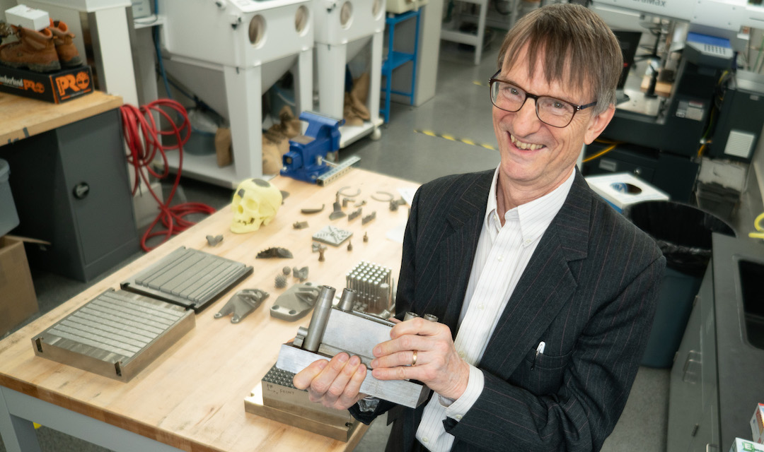 Tony Rollett in a lab with a table of 3d printed metal objects on the table next to him