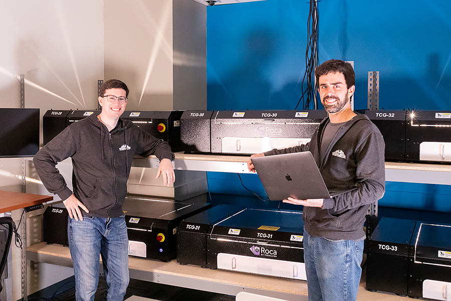 Two smiling men standing in front of what looks like boxes on shelves (but is actually the sorting robots). One man is holding a robot.