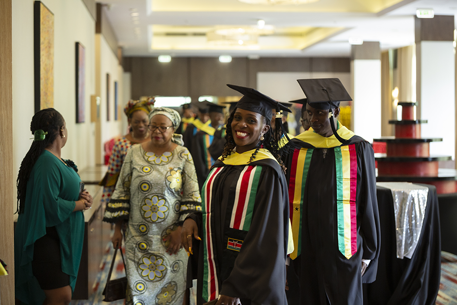 A CMU-Africa student poses in a hallway while others in graduation garb file out behind her