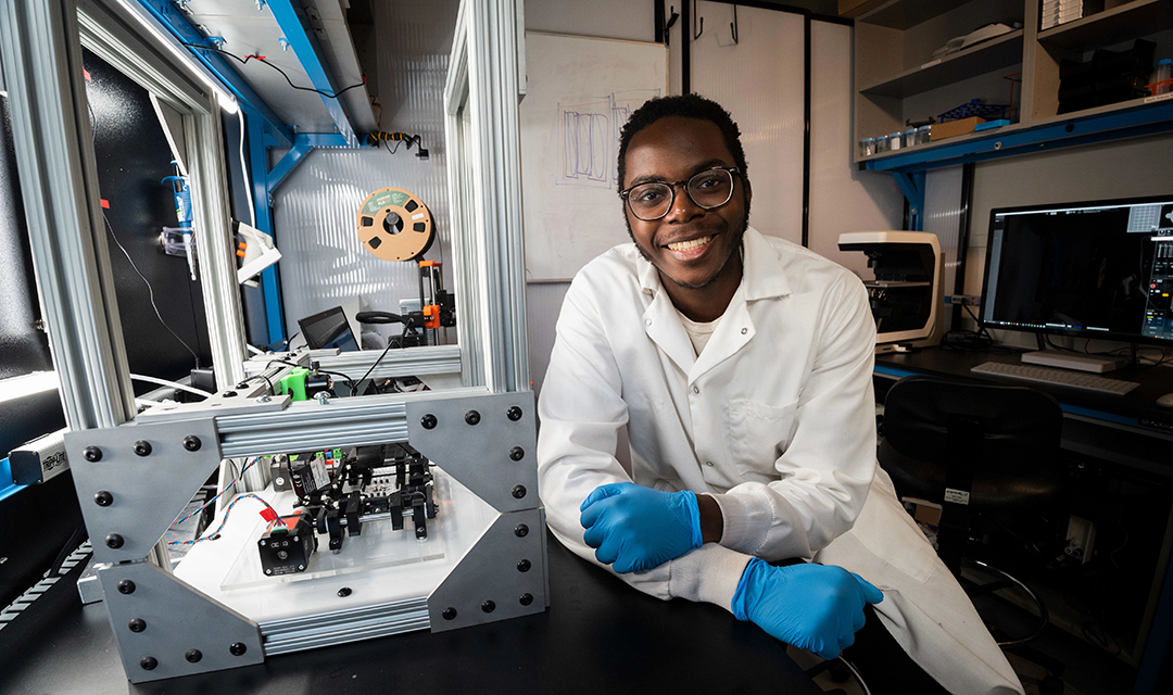 Lameck Beni in a lab next to a large glass box full of robotics equipment