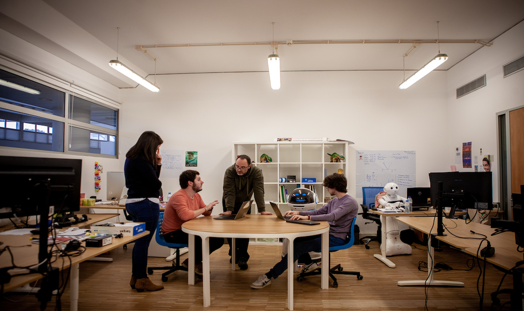 Four people chatting around a circular table in a room with cubbies in the back and other creative items