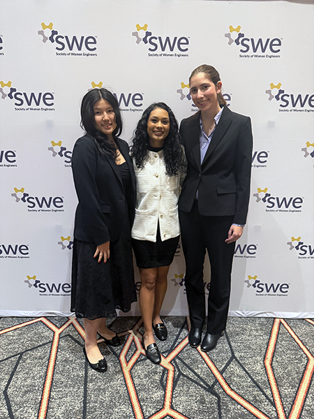 Three women standing in front of a wall with the SWE logo on it multiple times