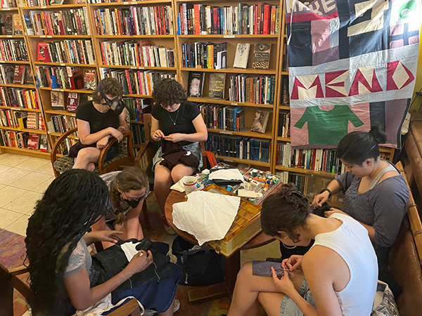 Six students in a library working on their mending project with a quilt hanging behind them that says "Mend"