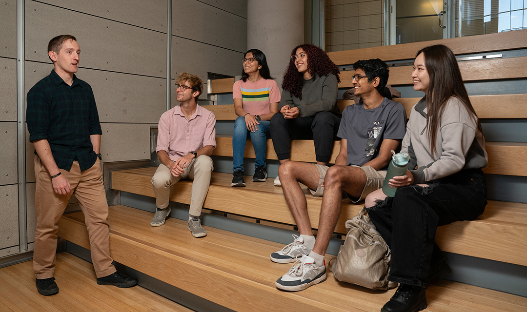 A student stands and talks while other students, who are sitting on benches, listen to him