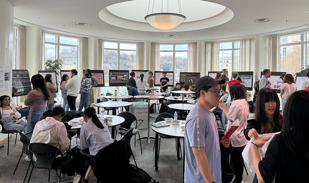 A room full of students collected around tables with posters for presentation around the edges of the room