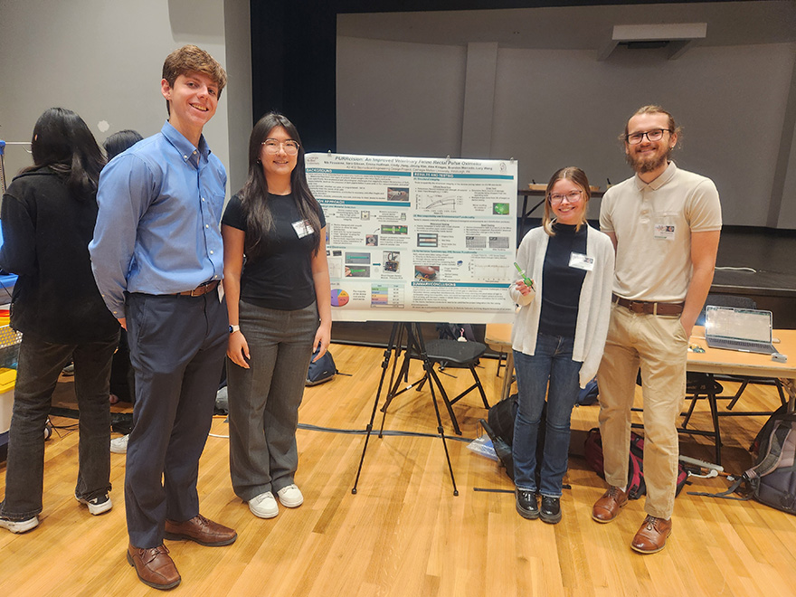 Four students wearing business casual attire stand near an academic poster. One student is holding a green oximeter prototype.