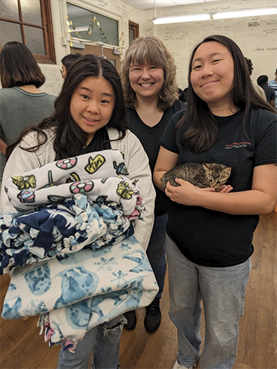 A young woman on the left holds soft blankets. A young woman on the right holds a small brown kitten. An older woman stands behind them smiling.