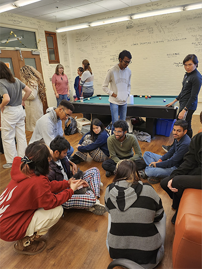 A group of students sit and stand in a lounge room with writing on the white walls, couches, and a pool table.