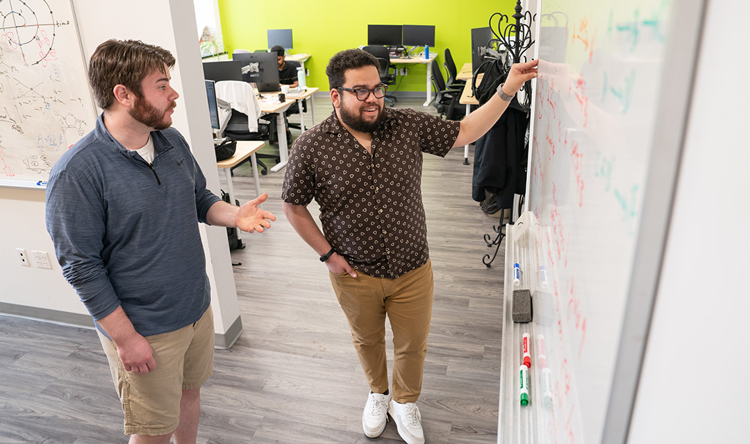 Gabe Gomes points at equations on a whiteboard while Robert MacKnight listens