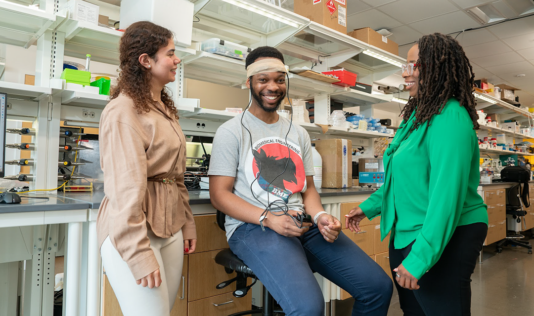 A young man sitting in a lab chair with sensors on his head is smiling, interacting with two women in a research laboratory setting.