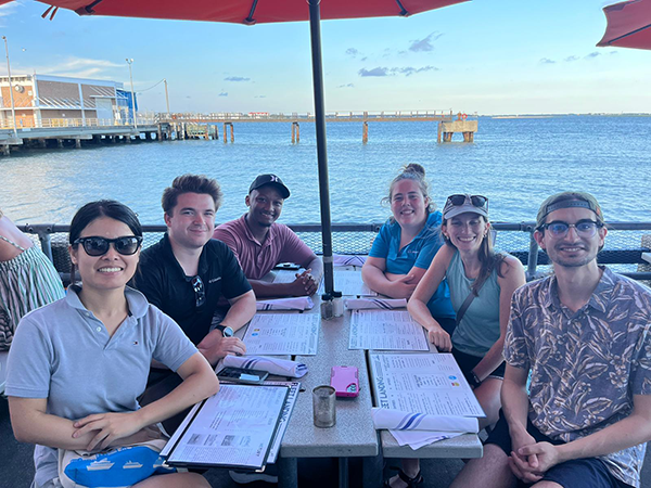 A group of six people at a restaurant table with the ocean behind them