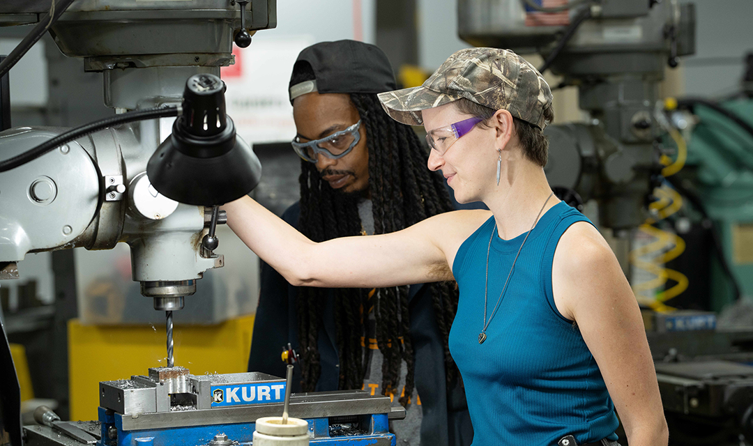A woman in a camouflage cap operates a milling machine while a man observes, both engaged in a manufacturing workshop.