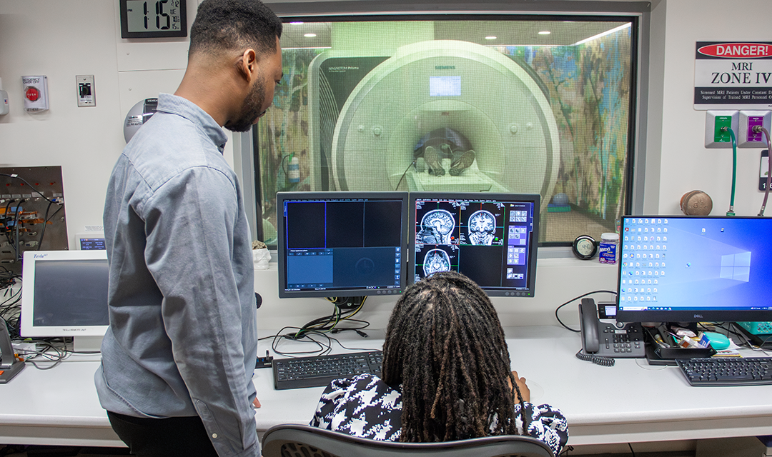 A technician and a patient monitor are visible in a medical imaging room, with the technician observing the MRI machine and the patient being scanned.