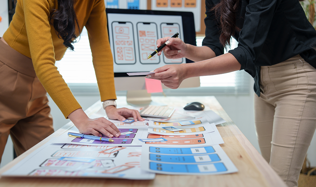 Two designers collaborate on a project, reviewing colorful wireframes and sketches on a table while one points to a design on a card.