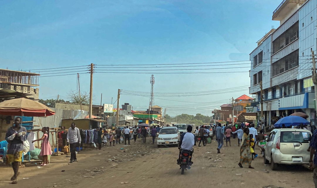 Busy street market on a dusty road with pedestrians, cars, and a motorbike, lined with shops and power lines under a clear blue sky.