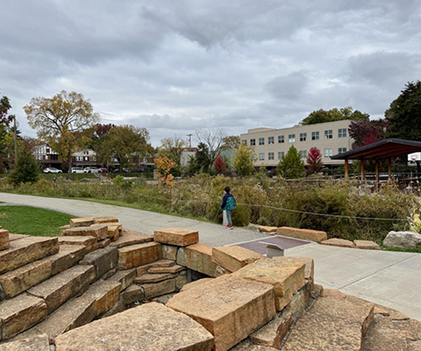 A student walking in front of a rock formation (the rocks are all even sizes). There is a building in the background.