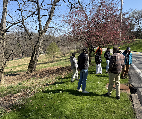 The same group of seven people that are in the second image in this slideshow. They are all outside in a park wearing coats standing near a road.