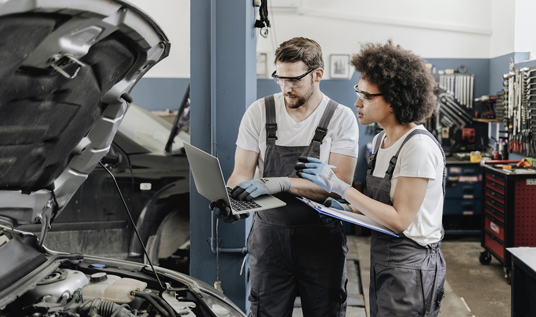 A man holding a laptop next to a woman with a clipboard, both looking under the hood of a car