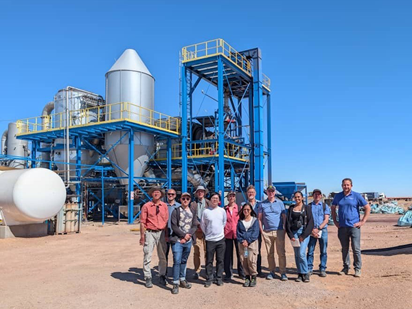 Group of people posing outdoors in front of a large industrial processing plant with blue steel framework and metal silos under a clear blue sky.