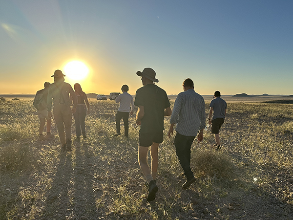Group of people walking across a grassy field at sunset, silhouetted against the low sun with long shadows.