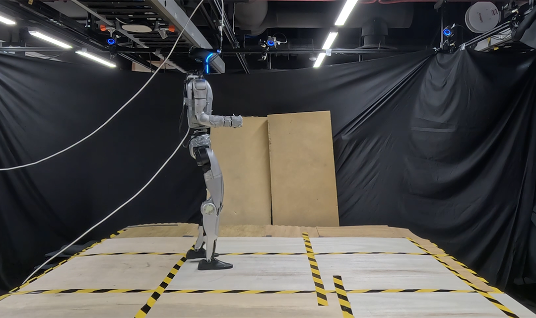 Humanoid robot standing on a taped test platform in a lab, surrounded by black curtains, overhead cameras, and two upright plywood panels.