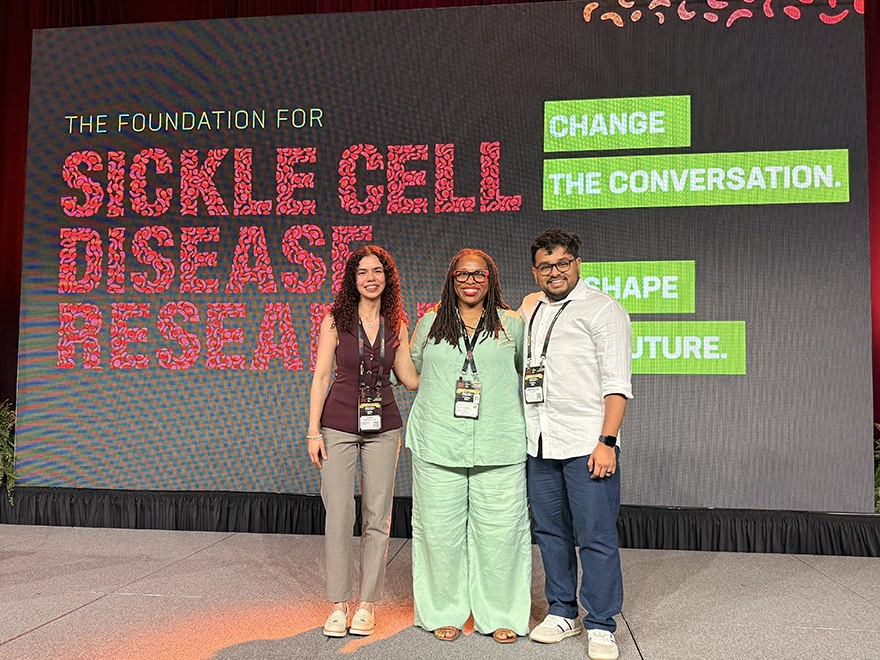 Three people stand on a stage in front of a screen that reads: Sickle Cell Disease Research