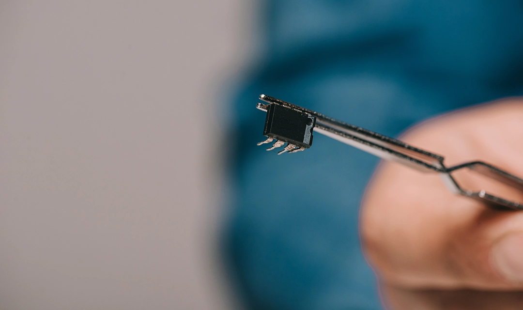 Close-up of tweezers holding a small integrated circuit chip with metal pins.