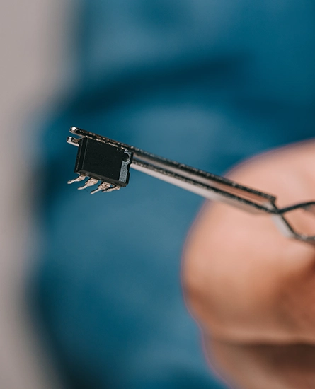 Close-up of tweezers holding a small integrated circuit chip with metal pins.
