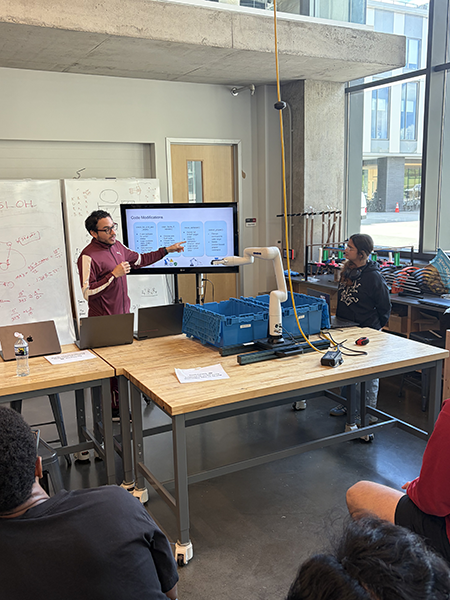 Student gestures toward a presentation slide on a large monitor while others listen in a workshop-style classroom with worktables and equipment.