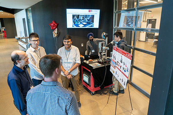 In the foreground, four people talk around a poster. In the background, two researchers demo their work.