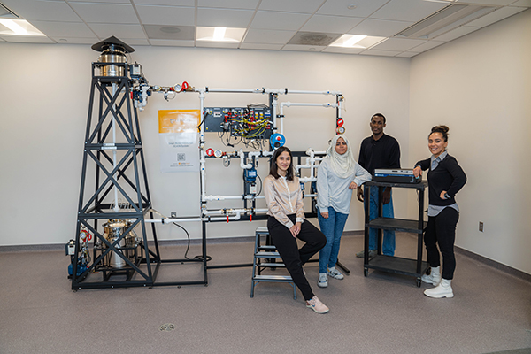 Four students pose in a lab beside a large piping and control-system training rig.